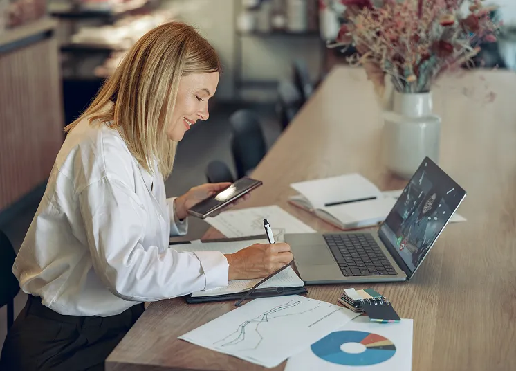 A woman sits at a table working on a laptop, taking notes on outsource CFO services in her notebook with charts and graphs nearby. She is smiling and holding a phone, participating in a video call.