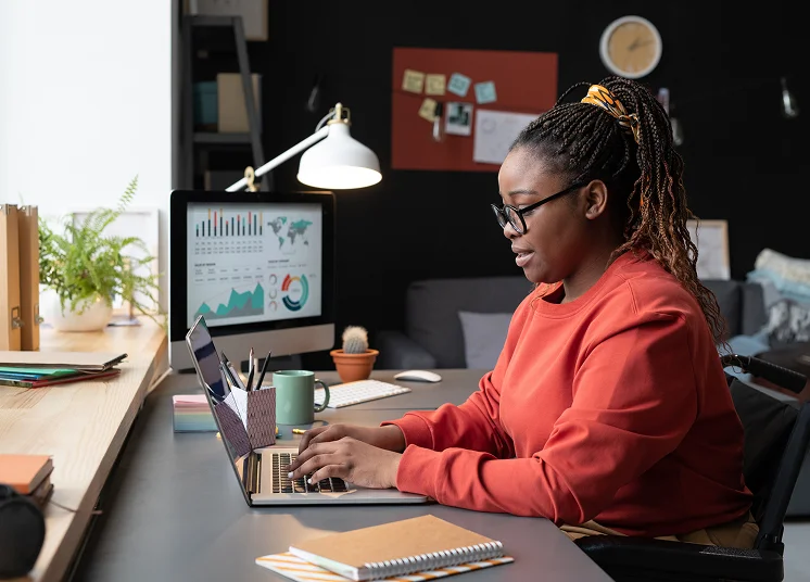 A woman wearing glasses and an orange sweatshirt works on a laptop at a desk. A desktop monitor displays graphs and charts. The workspace includes a notebook, cup, plant, and lamp, with a cozy room in the background.