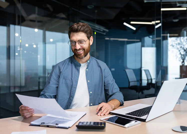 A smiling man with glasses sits at a desk in a modern office, holding a paper—ready to discuss CFO consulting services. A laptop, calculator, clipboard, and documents are on the desk. Glass walls and office chairs are in the background.