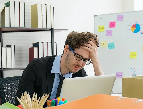 A man in a suit and glasses sits at a desk, looking stressed with his hand on his forehead while staring at a laptop. Office supplies, binders, and a whiteboard with notes and charts are visible in the background.