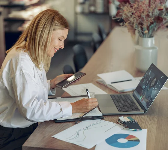 A woman sits at a desk working on a laptop, holding a phone and writing in a notebook. Papers about CFO consulting services with charts and graphs are spread out, and a vase with dried flowers is on the table.