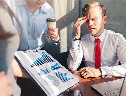 A stressed businessman in a white shirt and red tie sits at a desk with a laptop, rubbing his forehead, while colleagues show him a clipboard with charts and offer him a coffee.