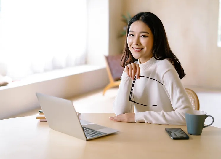 A woman sits at a desk with a laptop, holding glasses and smiling, ready to discuss outsource CFO services. A smartphone, cup, and notebook rest on the table as bright window light fills the room.