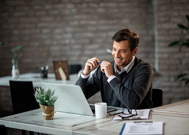 A man in a sweater sits at a desk with a laptop, smiling while holding a pen—perhaps reviewing financials as he considers outsource CFO services. Papers, a plant, and a coffee cup rest on the desk in the softly lit modern office.