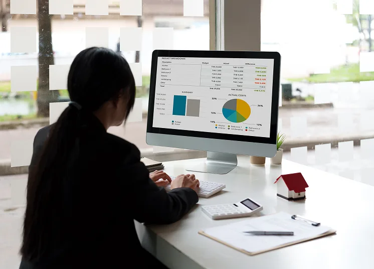 A person in business attire is sitting at a desk, working on a computer displaying charts and financial data. Nearby are a calculator, notepad, pen, and a small model house. Large windows are in the background.