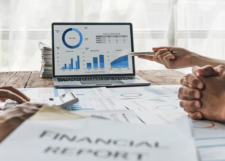 A laptop displaying financial graphs and charts sits on a table with printed reports labeled "FINANCIAL REPORT." Two people review the documents, one holding a pen and another with hands clasped.