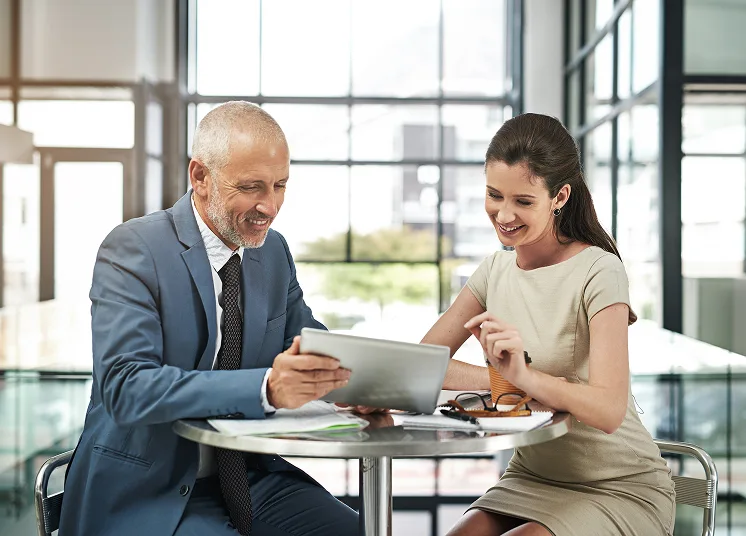 Two business professionals sitting at a table in a modern office, smiling while looking at a tablet. A notepad, pen, and a glass of juice are on the table. The background shows large windows with natural light.