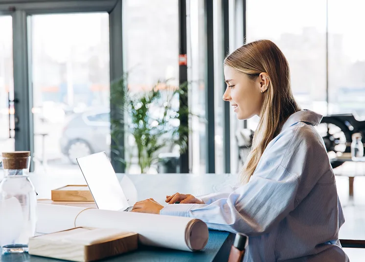 A woman with long blonde hair sits at a desk by large windows, working on a laptop. Rolled-up papers, a notebook, and a water bottle are on the table. Bright natural light fills the modern workspace.