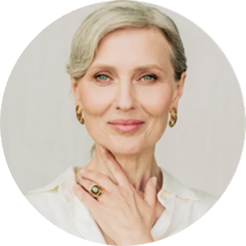 Portrait of a mature woman with light hair pulled back, wearing gold hoop earrings and a white blouse, gently touching her neck with one hand. She has a calm, confident expression.