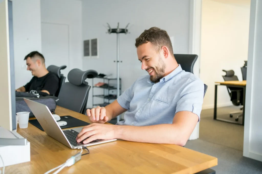 Two men sitting at desks in a modern office. One man in the foreground is smiling and working on a laptop, utilizing the best outsourced CFO services, while the other man in the background is also smiling and looking at his computer.