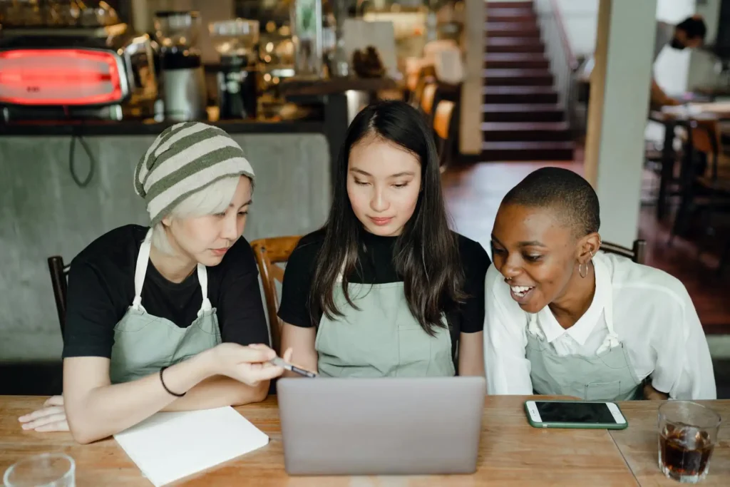 Three young women wearing aprons sit at a cafe table, focused on a laptop. As one points at the screen and another smiles, they discuss plans to hire virtual CFO services. A phone and drinks rest on the table, with the cozy cafe background visible.