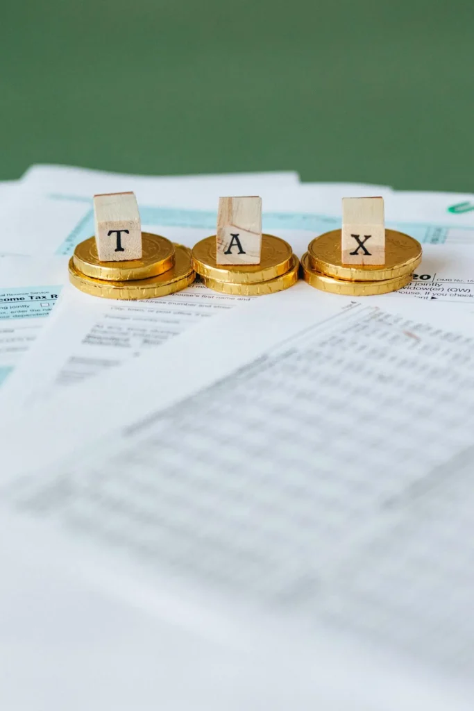 Scrabble tiles spelling "TAX" sit on stacks of gold coins, placed atop scattered tax documents—an image that highlights the benefits when you hire virtual CFO services for efficient financial management.