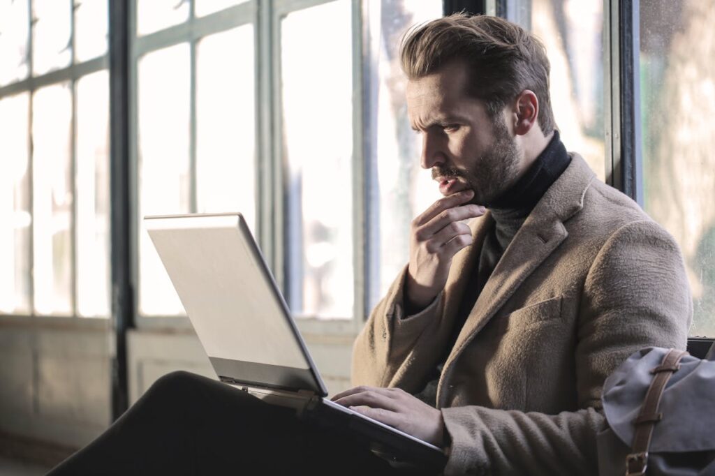 A man with a beard, wearing a tan coat and black turtleneck, sits indoors by large windows, looking thoughtfully at a laptop screen as he considers whether to hire virtual CFO services.