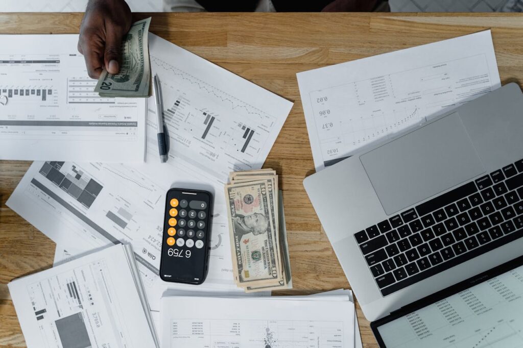 A person holds US dollar bills over a desk cluttered with financial documents, a pen, more cash, a smartphone displaying a calculator app, and an open laptop—perfectly illustrating why many business owners hire virtual CFO services.