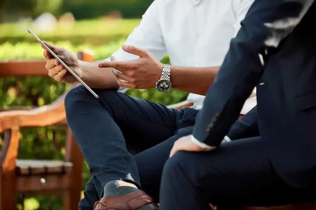 Two men in business attire sit outdoors on a bench. One man holds a tablet and gestures while discussing, while the other listens. Greenery and blurred background suggest a casual meeting in a garden or park.