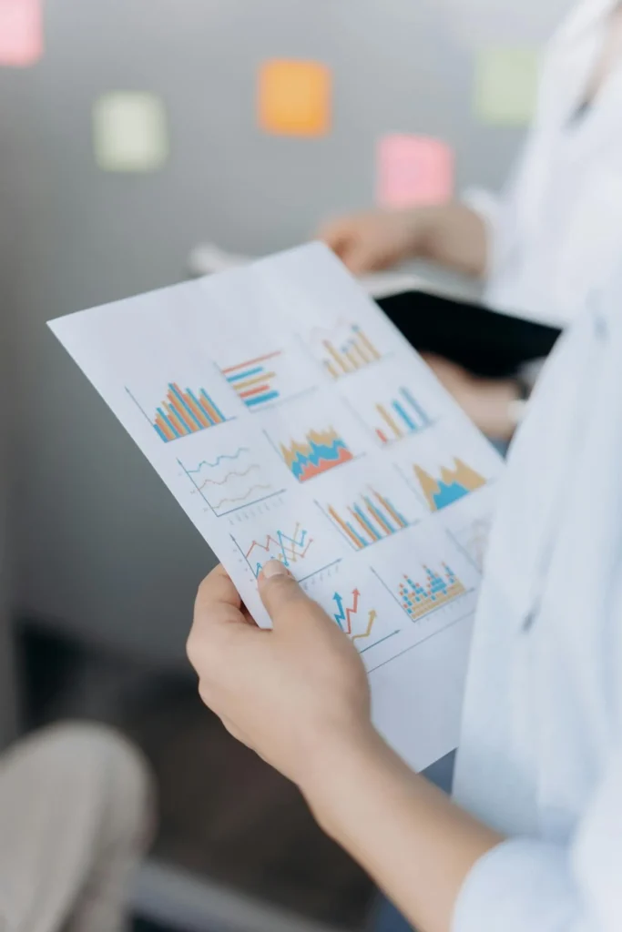 A person holding a sheet of paper displaying various colorful bar and line graphs, highlighting key insights from cfo outsourcing services, with blurred sticky notes and another person in the background.