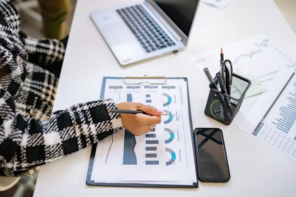 A person in a plaid jacket reviews charts and graphs on a clipboard at a desk with a laptop, phone, pens in a holder, printed financial documents, and materials related to CFO outsourcing services.