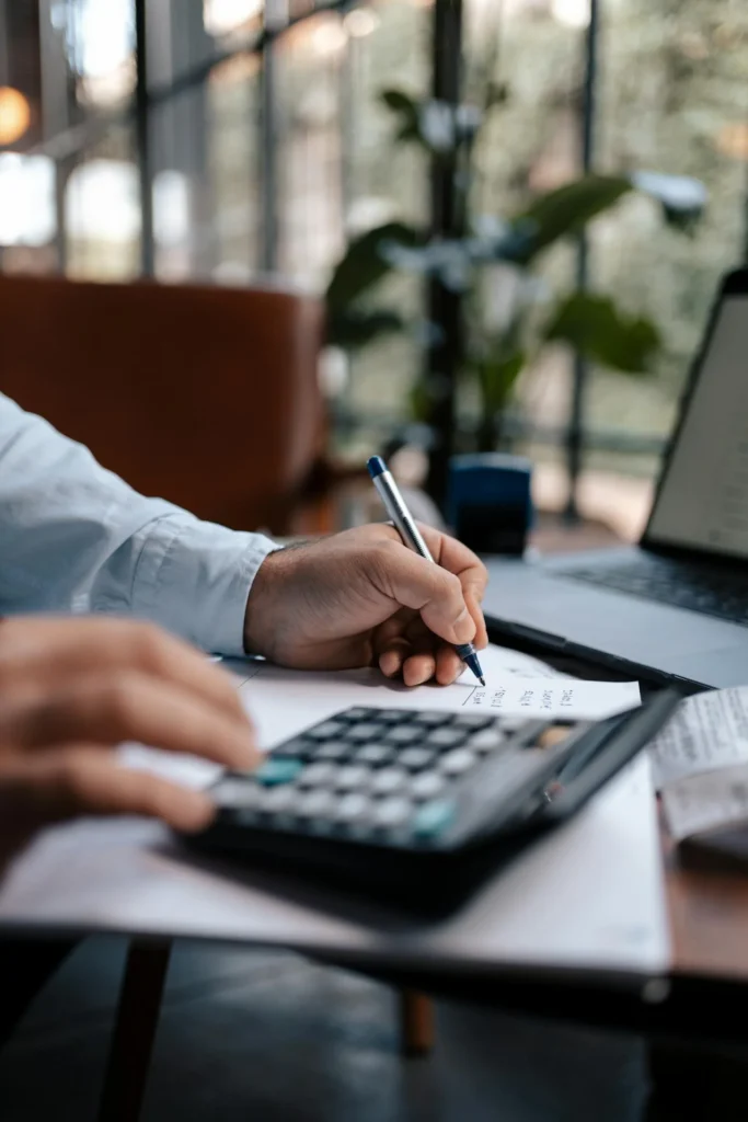 A person uses a calculator and writes on a piece of paper, with a laptop open nearby in a bright, modern workspace—perfect for managing finances or exploring CFO outsourcing services.