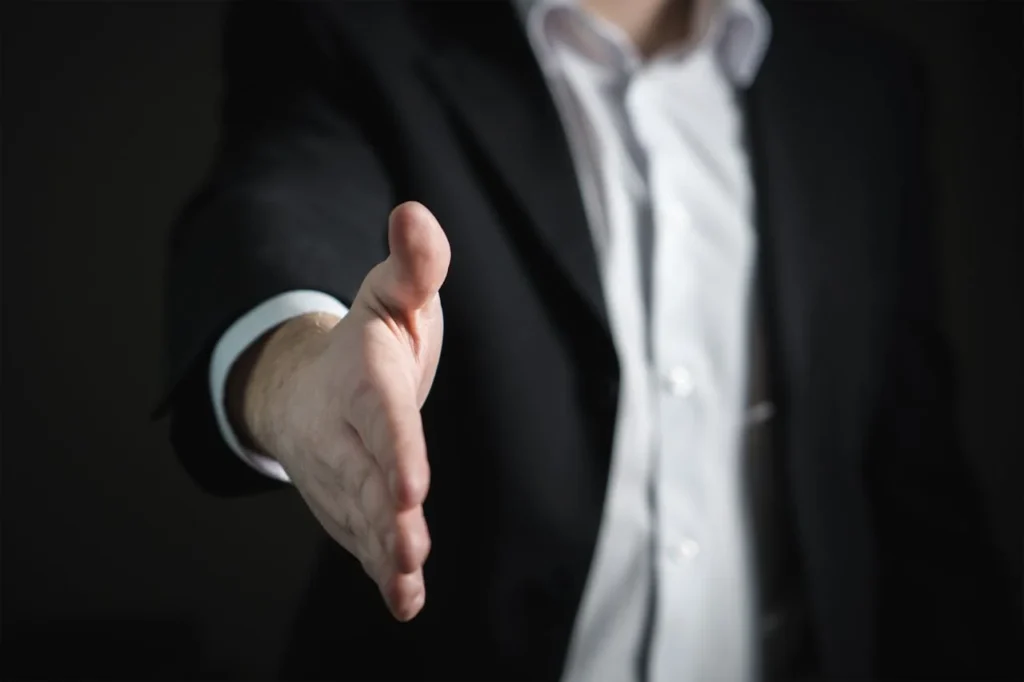 A person in a suit extends their right hand forward, offering a handshake. The dark background and focus on the gesture suggest a professional greeting or agreement, perfect for illustrating cfo outsourcing services.
