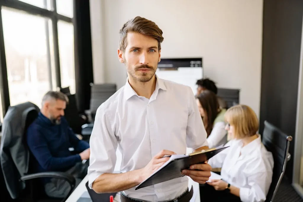 A man in a white shirt holds a clipboard and looks at the camera in a modern office, representing cfo outsourcing services, while four colleagues work and talk together in the background. Sunlight streams in through large windows.