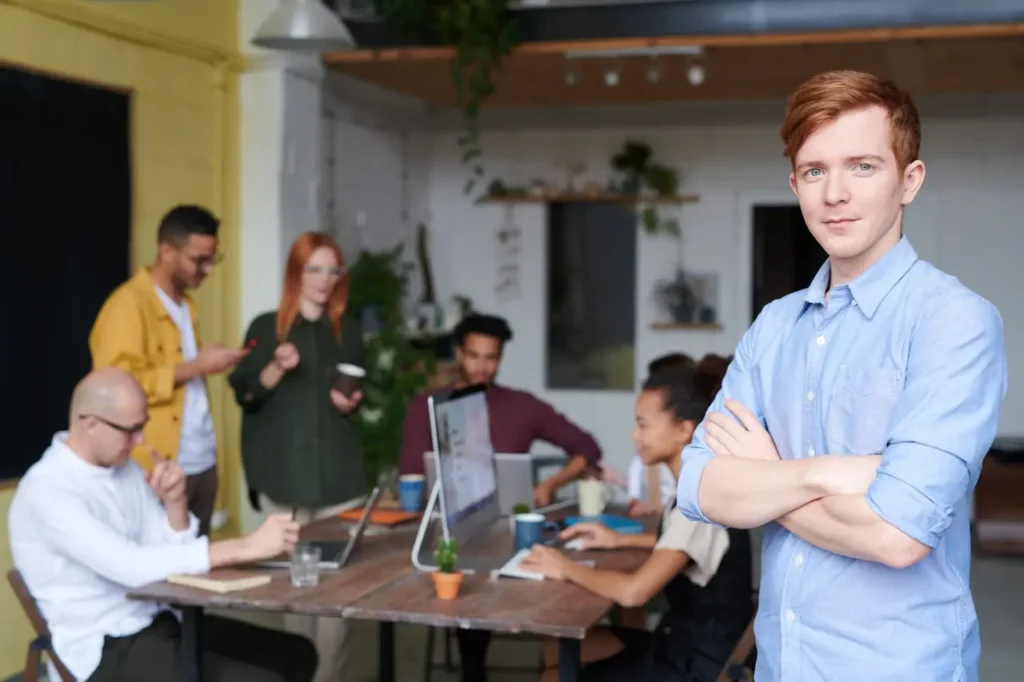 A young person with red hair stands confidently with arms crossed in the foreground, while a diverse group collaborates around a table with laptops and coffee in a modern office space, discussing innovative CFO outsourcing services.