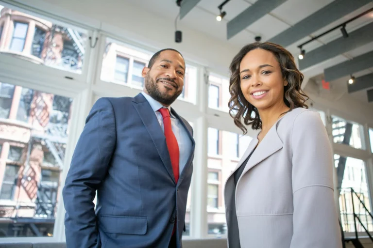 Two professionally dressed people, a man in a blue suit and red tie and a woman in a light gray blazer, smiling and standing in a modern office with large windows—illustrating the professionalism of top cfo outsourcing services.