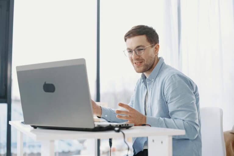 A man wearing glasses and a light blue shirt sits at a white desk, smiling and gesturing with his hands while participating in a video call on a laptop in a bright, modern room.