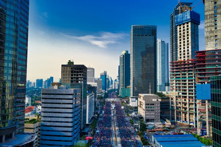 A wide city street in Jakarta, Indonesia, filled with people and bikes, is surrounded by tall modern skyscrapers under a clear blue sky, with some buildings still under construction.