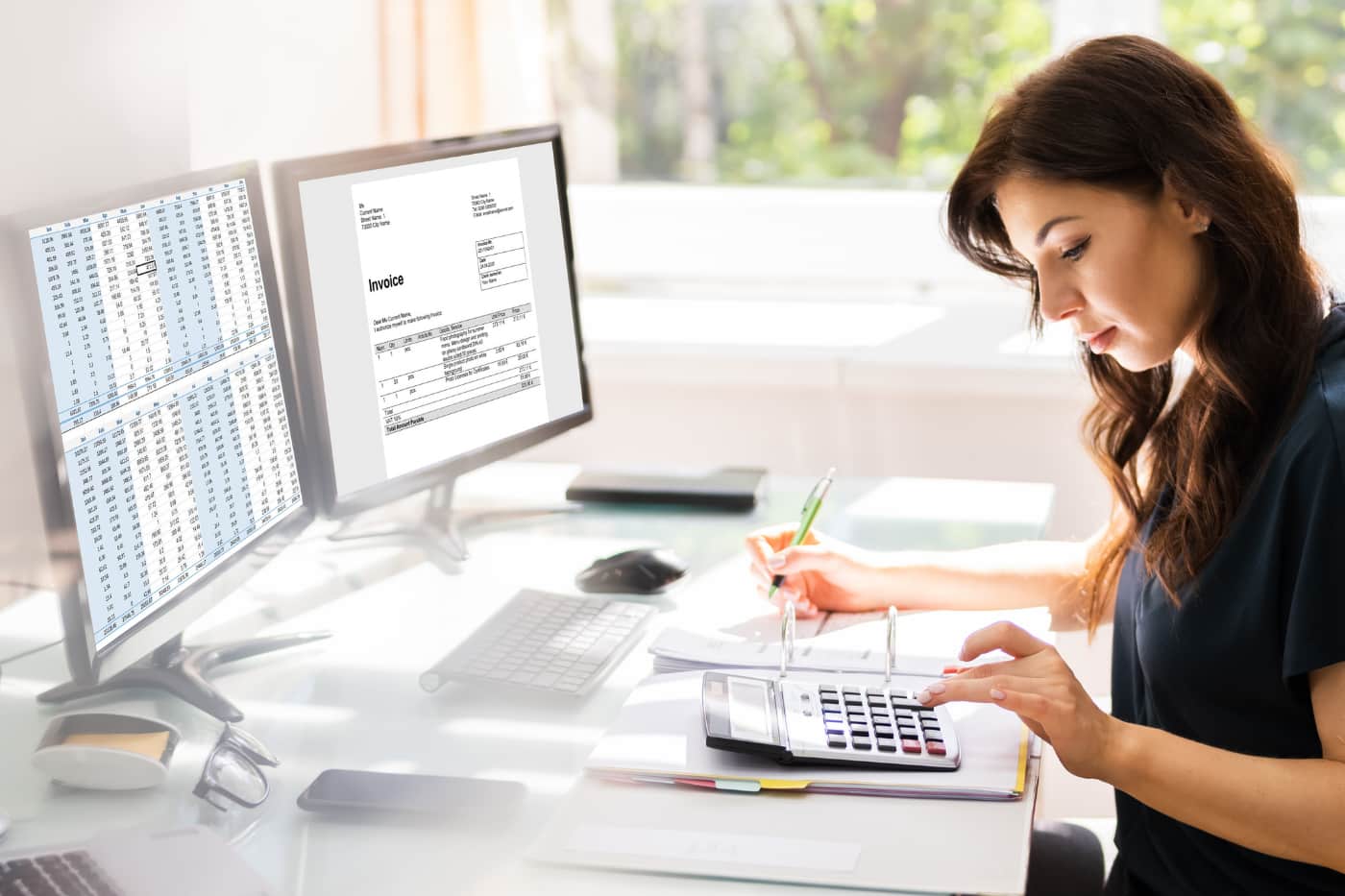 A woman sits at a desk using a calculator and writing in a notebook. She is working with documents, while two computer monitors display a spreadsheet and an invoice. Sunlight streams through a window nearby.