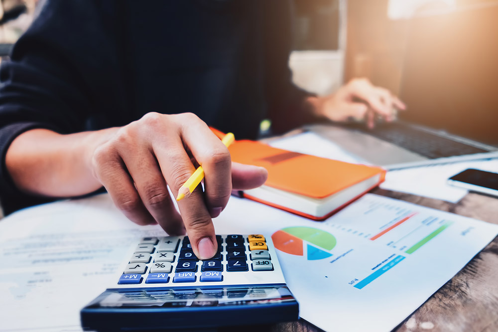 A person uses a calculator while holding a pencil, with a notebook, laptop, and a financial chart with colorful pie graphs on a desk, suggesting they are working on finances or data analysis.