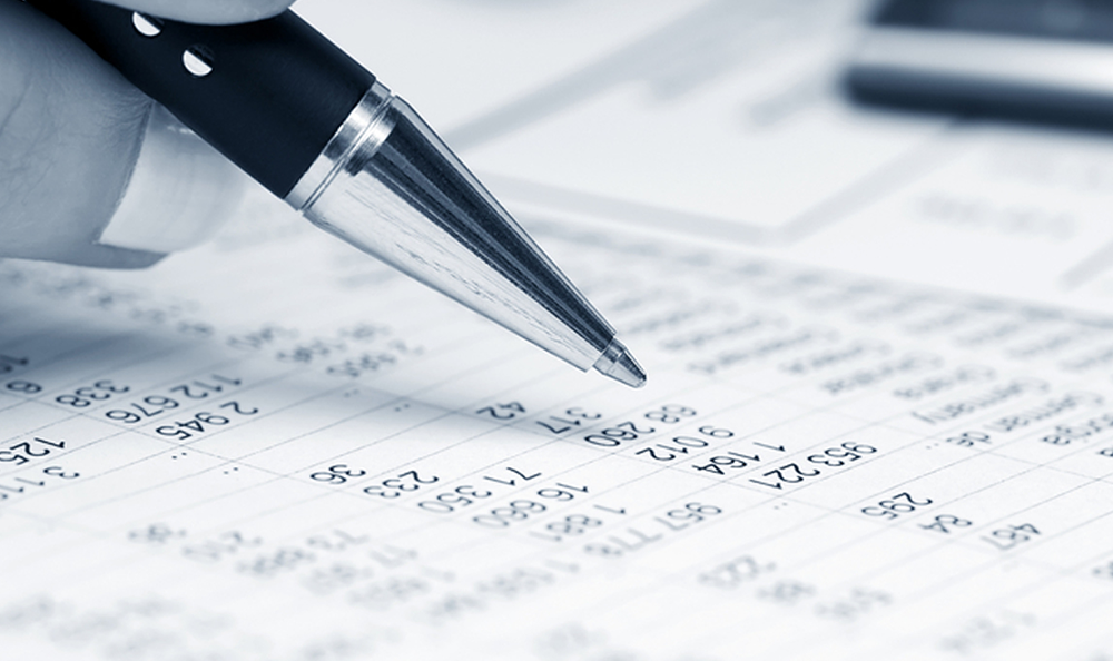 A close-up of a hand holding a pen and pointing at numbers on a financial spreadsheet, with blurred documents and a calculator in the background.