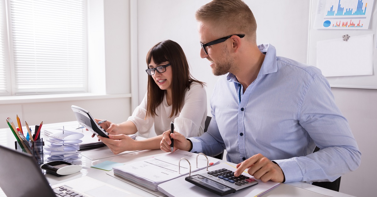 Two coworkers sit at a desk with paperwork, calculators, and binders. The woman smiles while showing a calculator to the man, who writes in a binder. Both wear glasses and work in a bright office with charts on the wall.