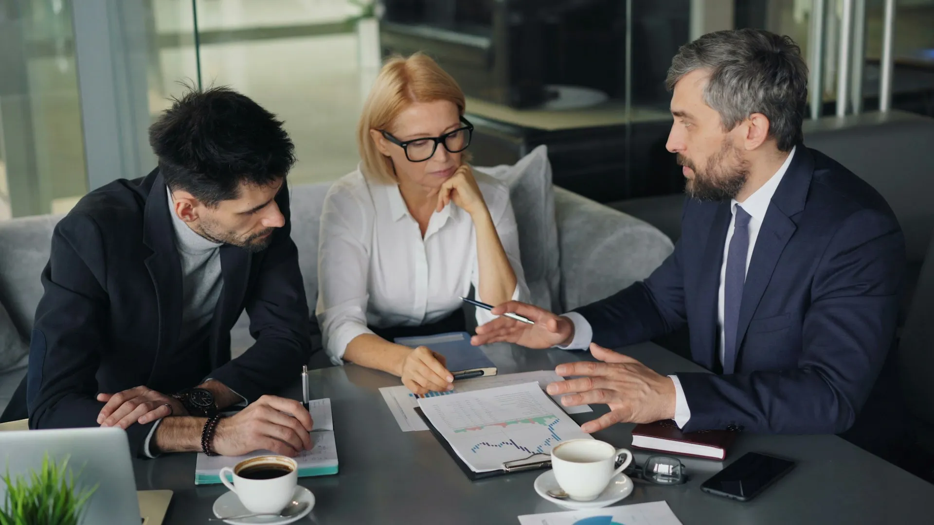 Three business professionals sit at a table with documents, charts, and coffee cups, engaged in a serious discussion. One man gestures while speaking, and the other two listen attentively, taking notes.